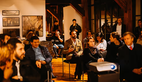 Group of people seated and standing in a warmly lit room, attentively listening to a speaker or presentation.