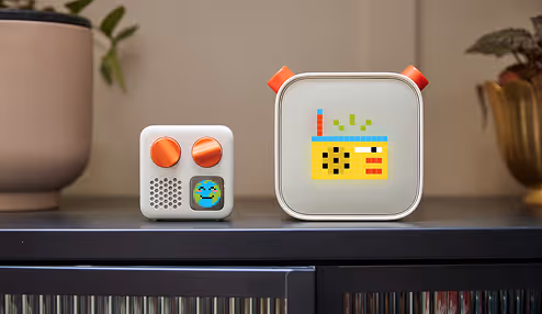 The Yoto player. Two compact modern electronic devices with knobs and pixelated displays placed on a black shelf with plants in the background.