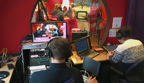 Two people wearing headphones operating sound and video equipment in a room with red walls and a circular window showing a group of people inside a studio.