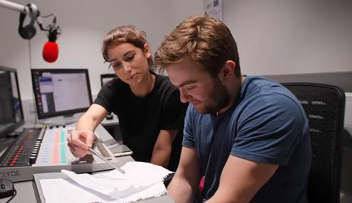 Two people collaborating over notes in a recording studio with microphone and computer screens.