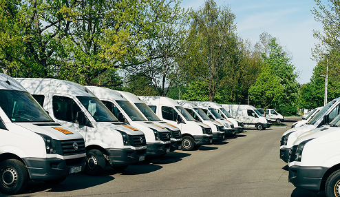 Row of white delivery vans parked along a road with green trees in the background.
