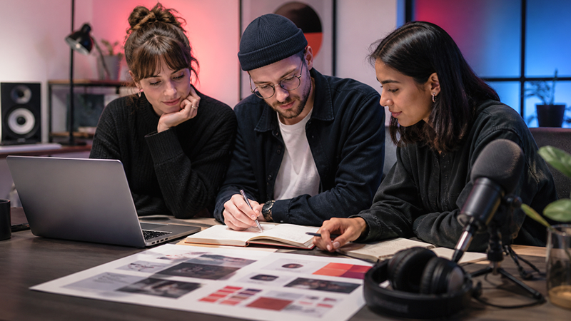 Three creative professionals collaborating around a desk with a laptop, design layouts, and notes, in a modern workspace.