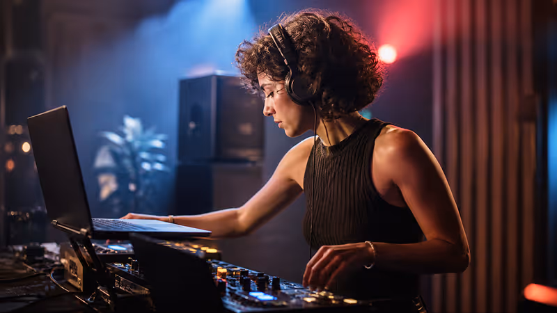Young woman DJ wearing headphones working on a laptop and mixer in a dimly lit club.