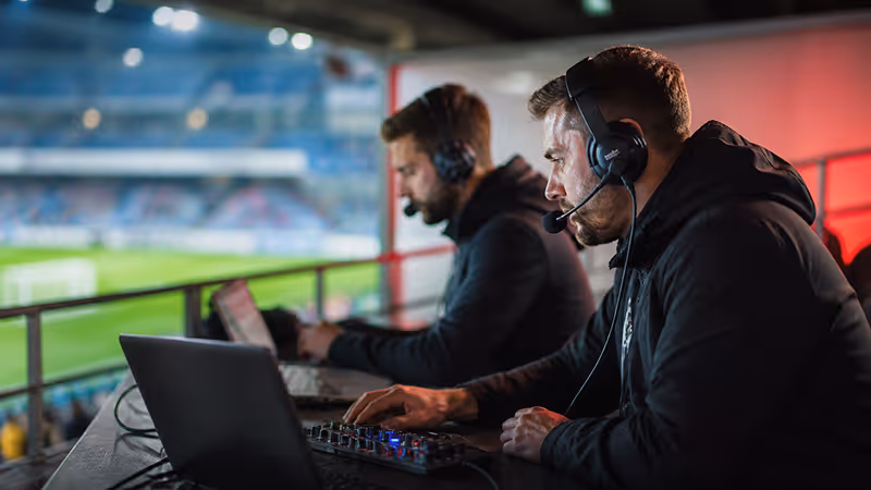 Two male sports broadcasters wearing headsets working on laptops and audio equipment in a stadium booth.