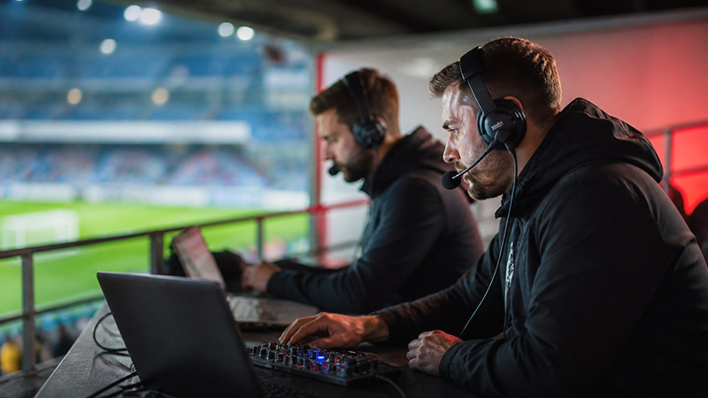 Two male sports broadcasters wearing headsets working on laptops and audio equipment in a stadium booth.