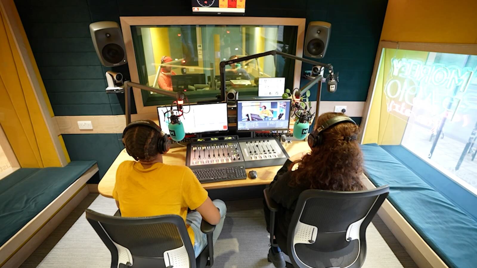 Two radio hosts wearing headphones sitting at a broadcast mixer with microphones in a studio.