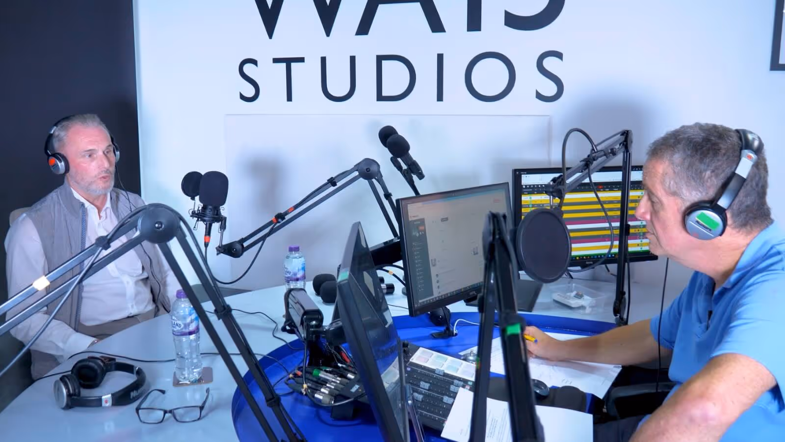 Two men wearing headphones speak into microphones during a radio show in a studio.