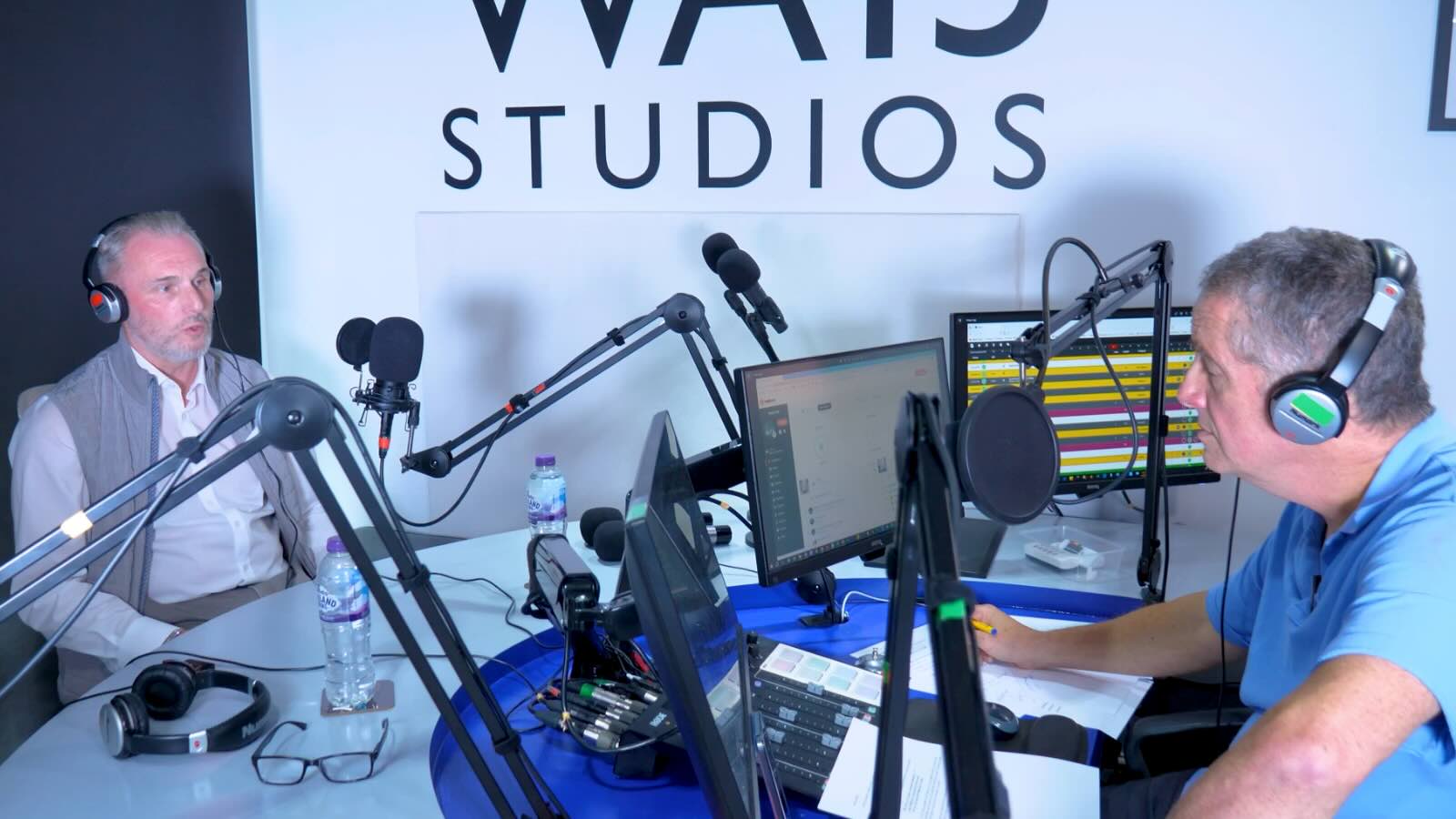 Two men wearing headphones speak into microphones during a radio show in a studio.