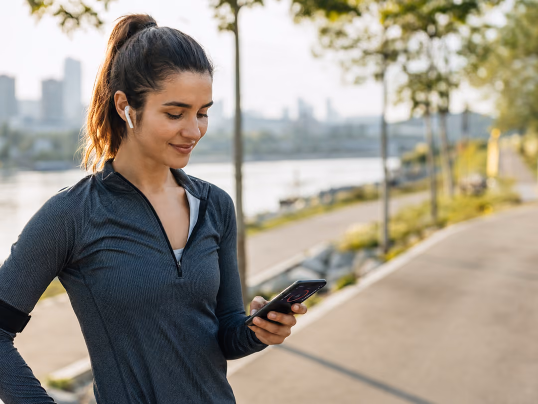 Woman in athletic wear with earbuds looking at her smartphone by a riverside path in the city.