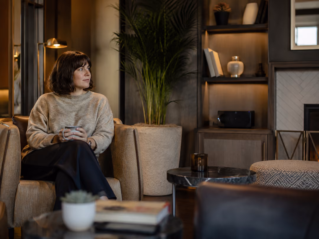Woman in beige sweater holding a cup, sitting in a cozy living room with plants and bookshelves.