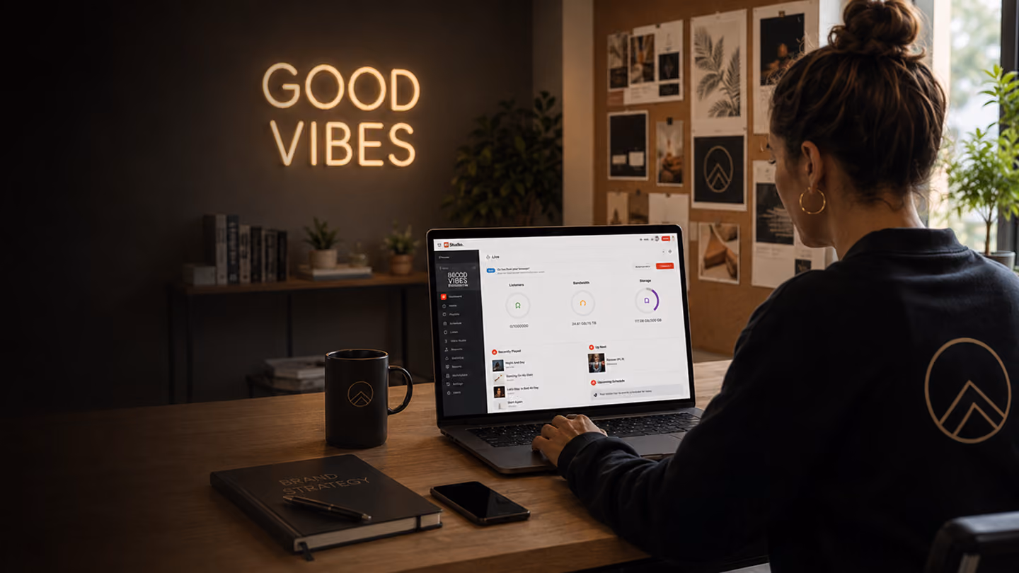 Woman working on laptop at desk with mug, phone, notebook, and 'Good Vibes' neon sign on wall.