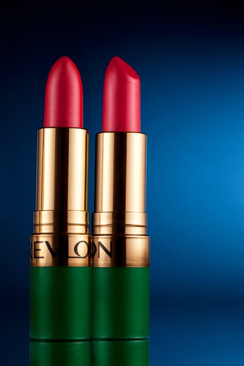 A product shot of two red lipsticks with green and gold base with blue gradient background. 