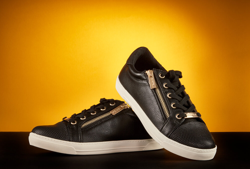 Product shot of black leather trainers with white soles and gold side zips on background of black and yellow. 