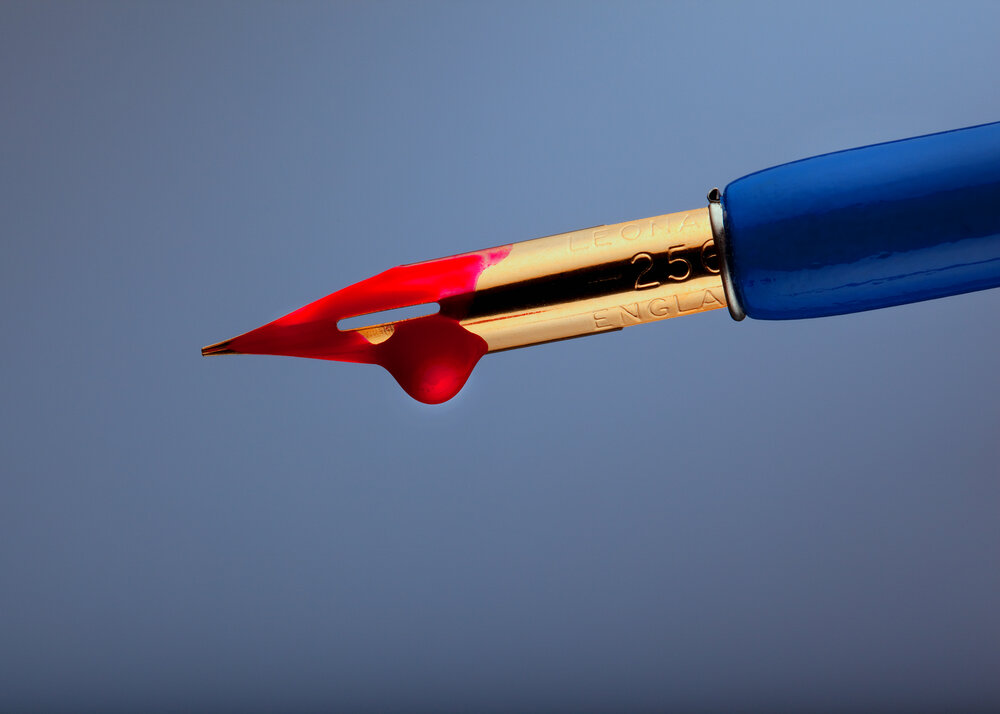 A product shot of a blue ink pen with golden nib. The nib is covered in blood red ink on a blue gradient background. 