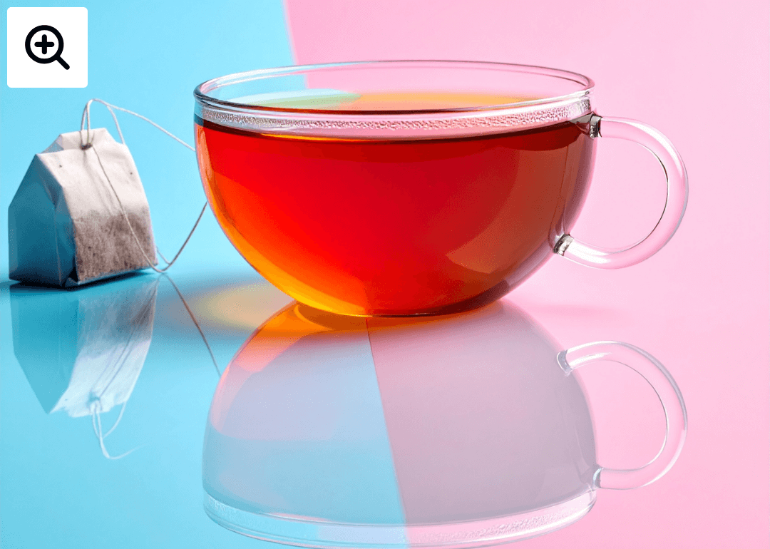 A glass tea cup with red tea in it on a reflective surface of pink and blue. A small house shaped tea bag sits alongside it. 
