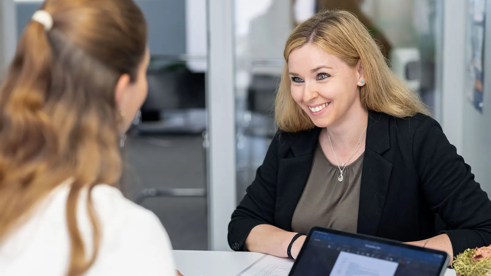 Zwei Personen sitzen sich gegenüber und führen ein Gespräch. Eine der Personen lächelt offen, während ein Laptop auf dem Tisch steht.