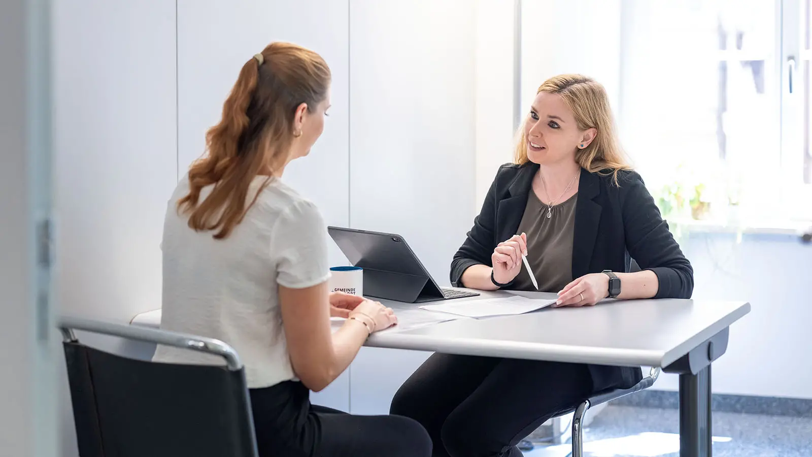 Zwei Personen sitzen sich an einem Tisch gegenüber und führen ein Gespräch. Eine Person erklärt etwas anhand von Unterlagen auf einem Tablet.