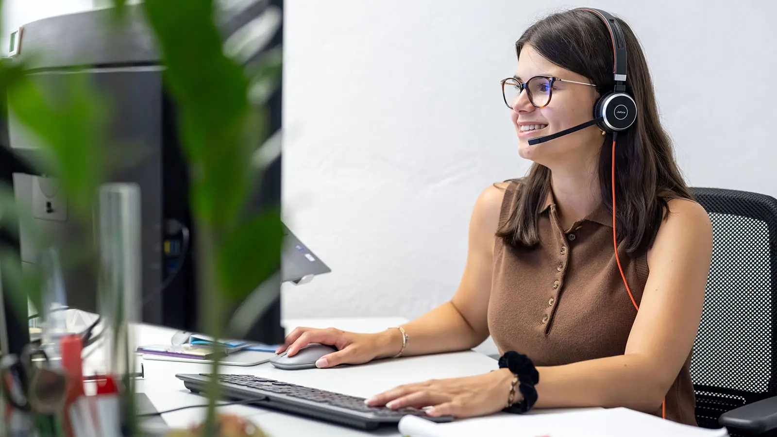 Eine Mitarbeiterin mit Headset sitzt am Schreibtisch und arbeitet am Computer. Sie lächelt, während sie mit einer anderen Person spricht.