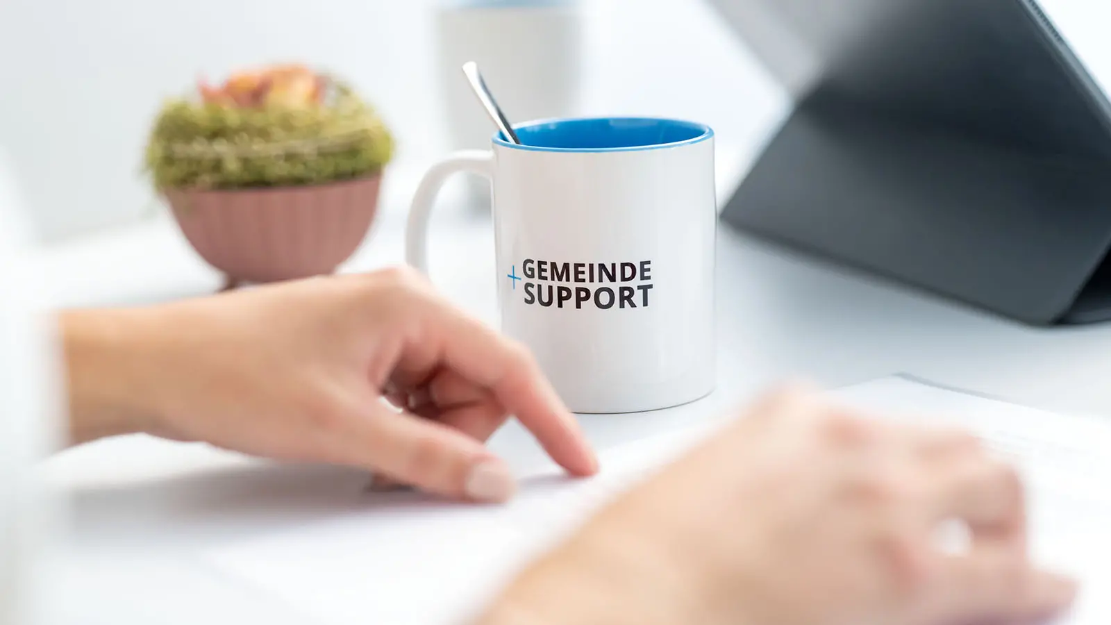 m Vordergrund liegen Unterlagen auf einem Tisch, im Hintergrund steht eine Tasse mit der Aufschrift „Gemeinde Support“. Eine Person zeigt auf ein Dokument.
