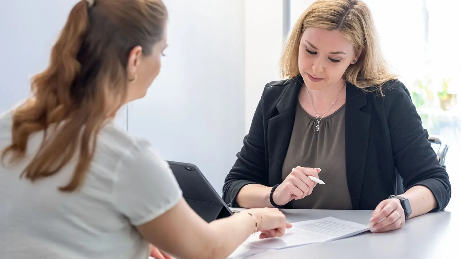 Zwei Personen sitzen sich an einem Tisch gegenüber und besprechen ein Dokument. Eine Person zeigt mit einem Stift auf das Papier, während die andere aufmerksam zuhört.
