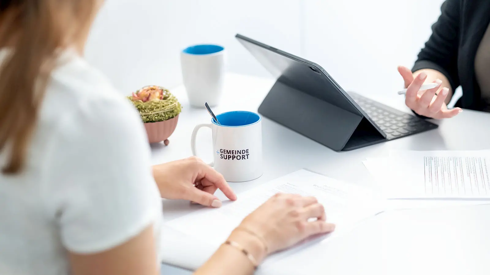Auf einem Tisch liegen Unterlagen, daneben stehen zwei Tassen. Eine Person zeigt auf ein Dokument, während eine andere ein Tablet mit Tastatur nutzt.