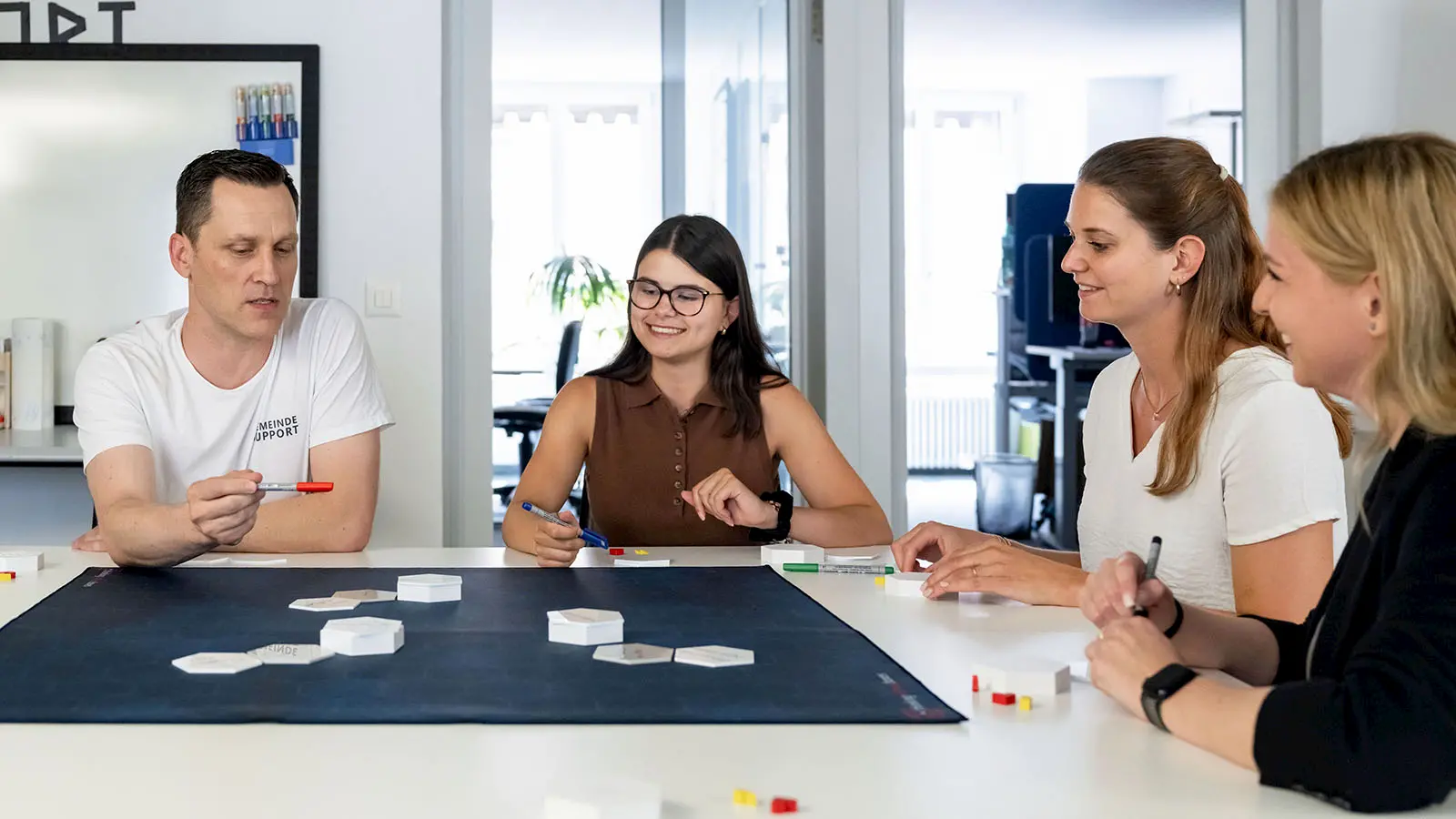 Vier Personen sitzen an einem Tisch und arbeiten gemeinsam mit Karten und Stiften an einer Aufgabenstellung. Sie tauschen sich konzentriert aus und entwickeln zusammen Lösungen.