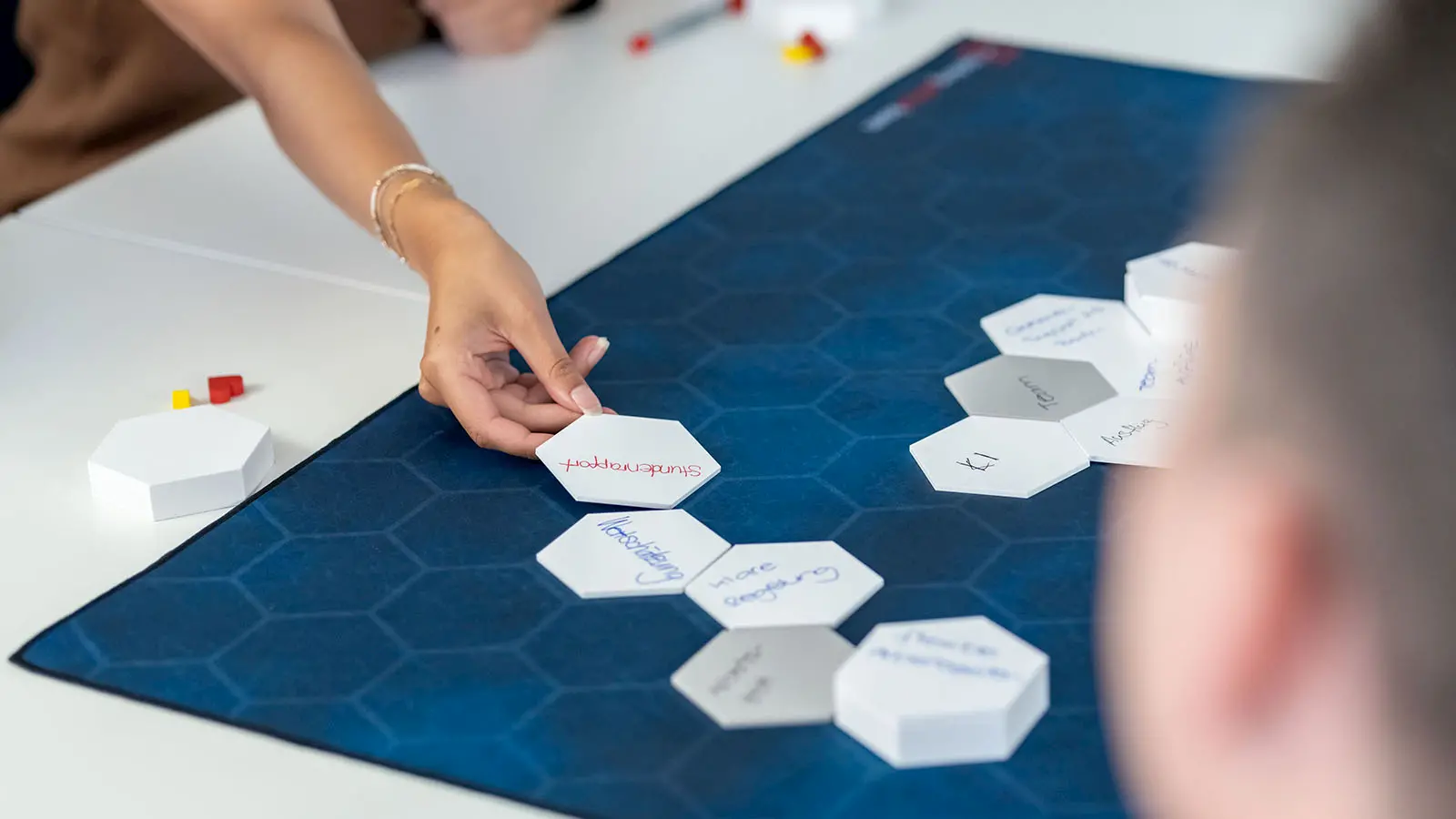 Eine Person legt eine beschriftete, sechseckige Karte auf eine blaue Arbeitsmatte, auf der weitere Karten mit handschriftlichen Notizen angeordnet sind.