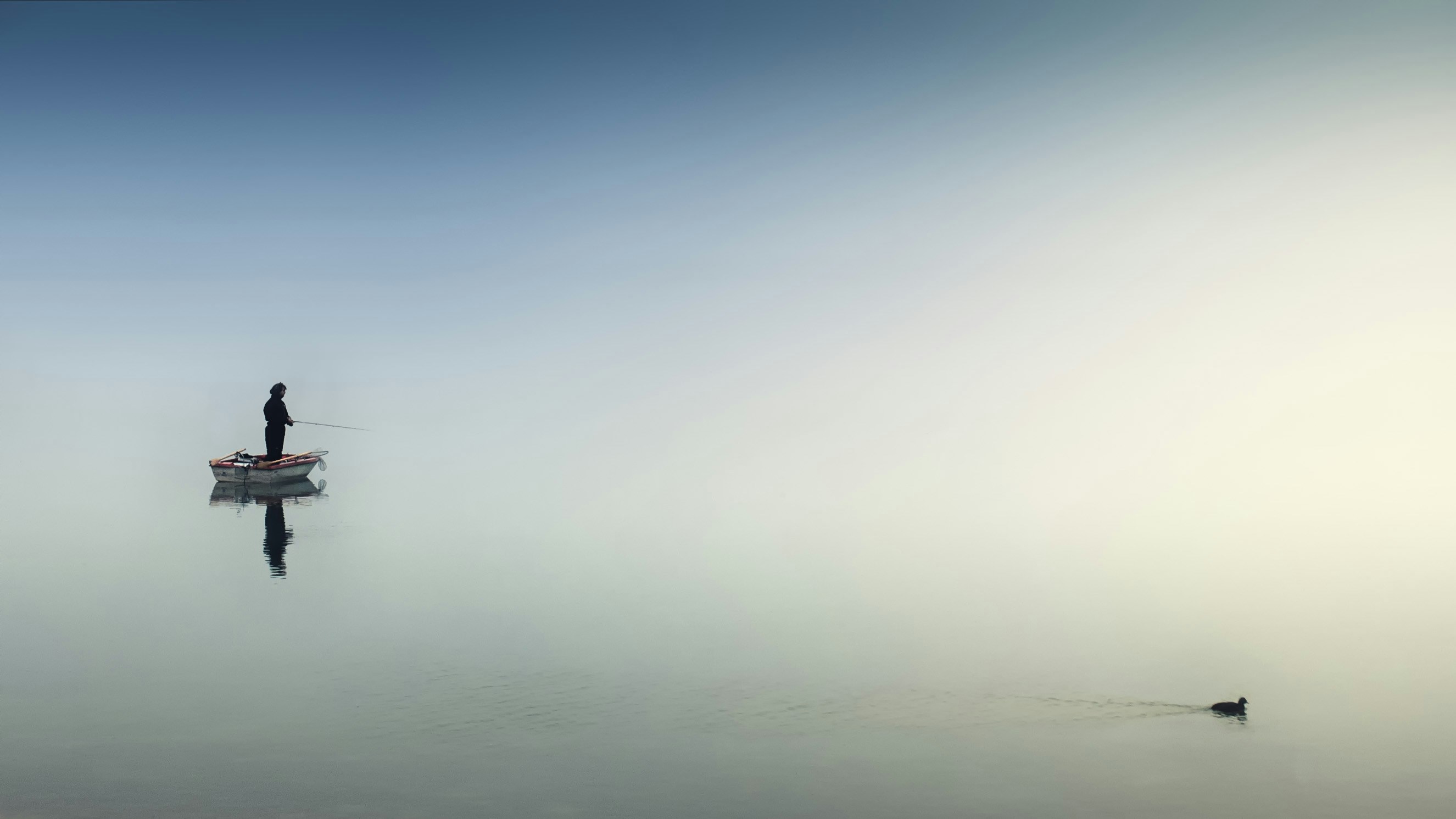 A man fishes on a boat while a duck swims by.