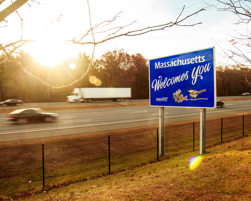 A blue roadside sign reading ‘Massachusetts Welcomes You’ stands beside a highway, with cars and trucks driving past and autumn trees in the background.