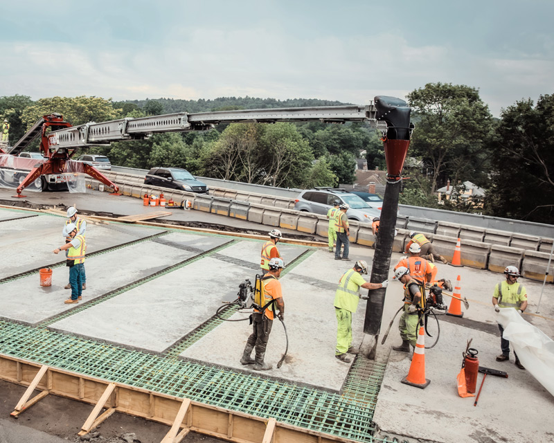 Construction workers wearing PPE pouring concrete on a bridge deck using a large concrete pump with an extended arm.