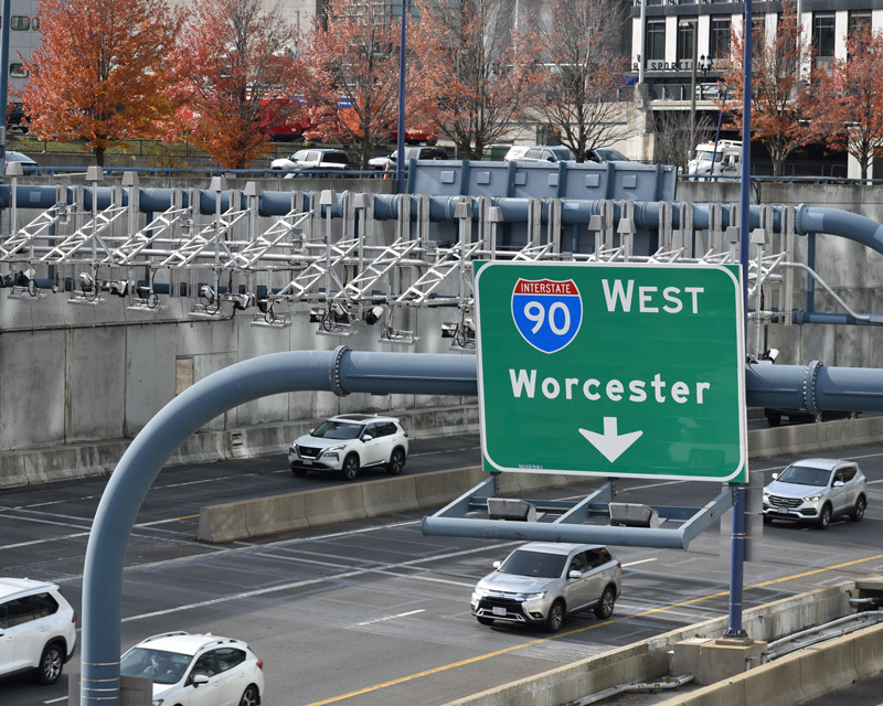 A sign over the MassPike indicating I-90 West towards Worcester, with several cars underneath and an electronic tolling gantry in the background.