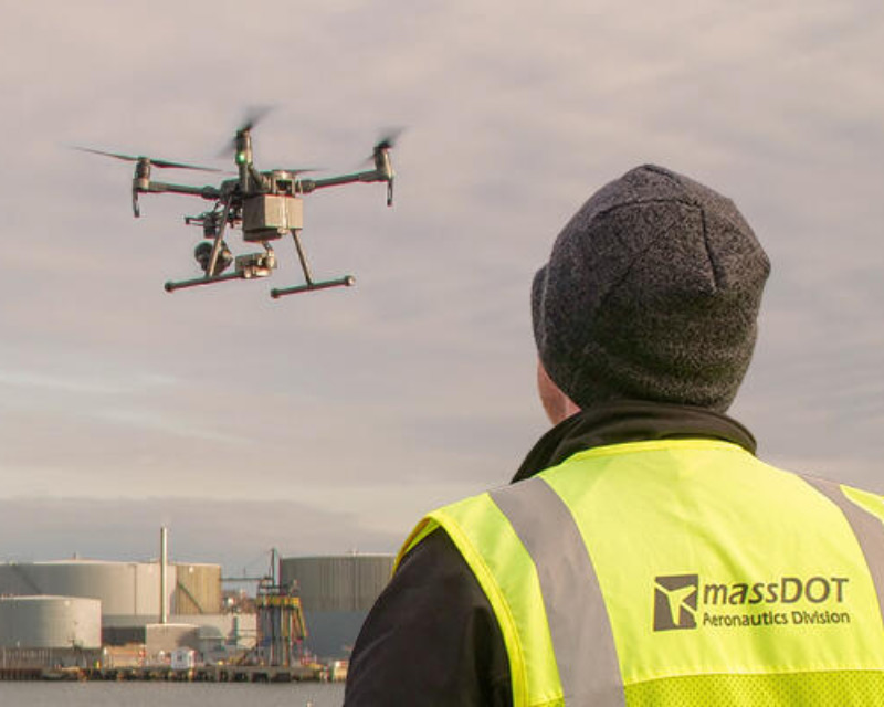 A MassDOT drone pilot in PPE operating a drone near industrial storage tanks under a cloudy sky.