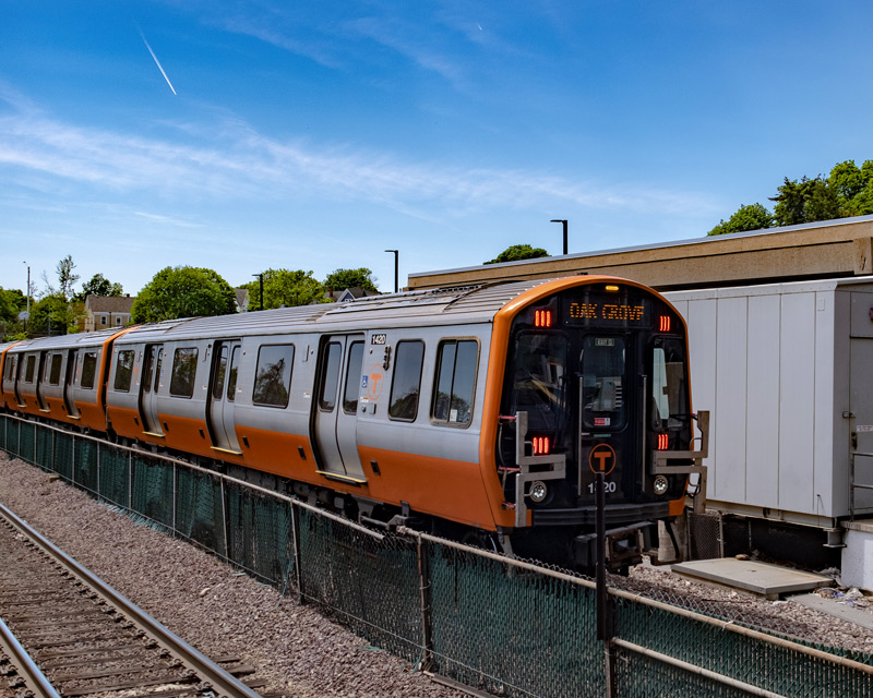 A new MBTA Orange Line train traveling past urban residential buildings under a clear sky.