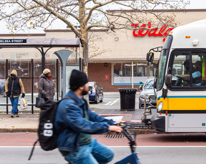 A cyclist riding a BlueBike rides past a MBTA bus stop on Broadway in Everett, with a MBTA bus pulling up and two passengers waiting for the bus.