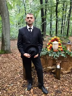 Mann in schwarzem Anzug hält schwarzes Hütchen, steht vor einem Urnengrab mit farbenfrohem Blumenkranz in einem Wald.