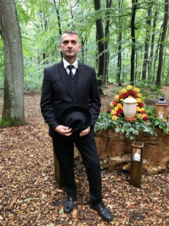 Mann in schwarzem Anzug hält schwarzes Hütchen, steht vor einem Urnengrab mit farbenfrohem Blumenkranz in einem Wald.