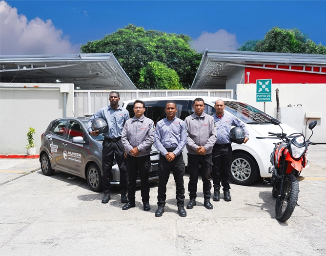 Five security guards in uniform standing in front of a gray car, a white van, and a red motorcycle in a parking lot.