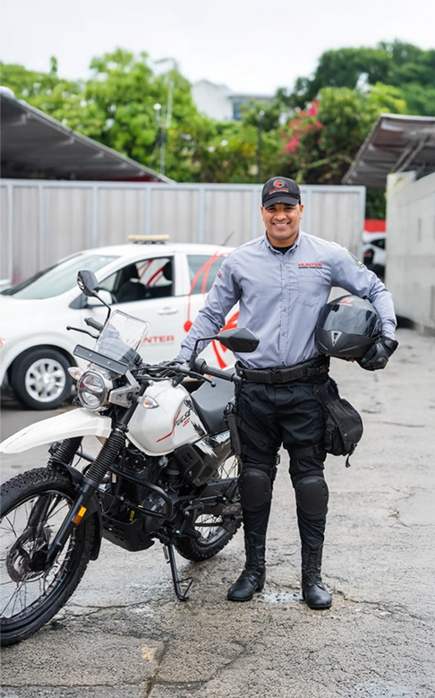 Smiling security officer in uniform standing beside a white police motorcycle holding a helmet in a parking area.