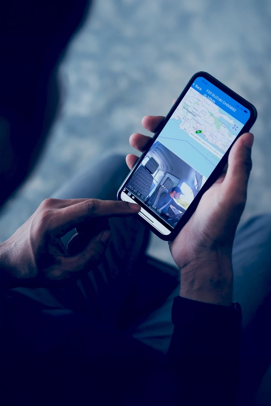 Person holding a smartphone displaying a vehicle tracking app with a map and live driver camera feed.