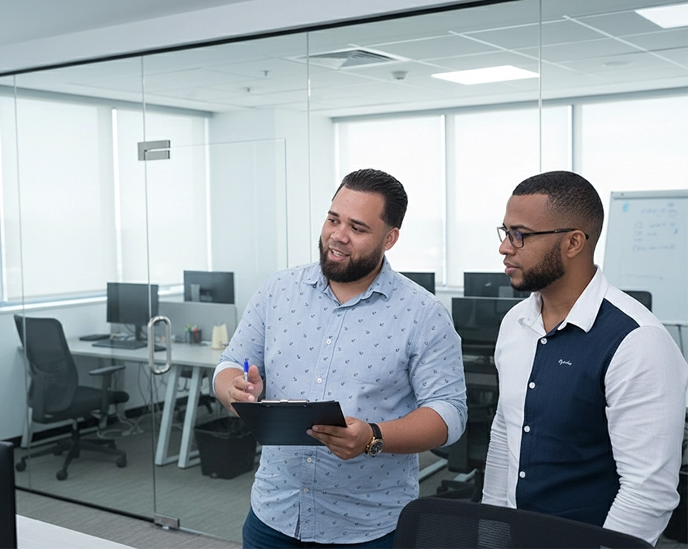 Two men standing and discussing in a modern office with glass walls, one holding a clipboard and pen.