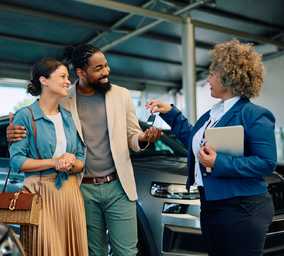 Car salesperson handing over keys to a smiling couple standing in front of a vehicle inside a dealership.