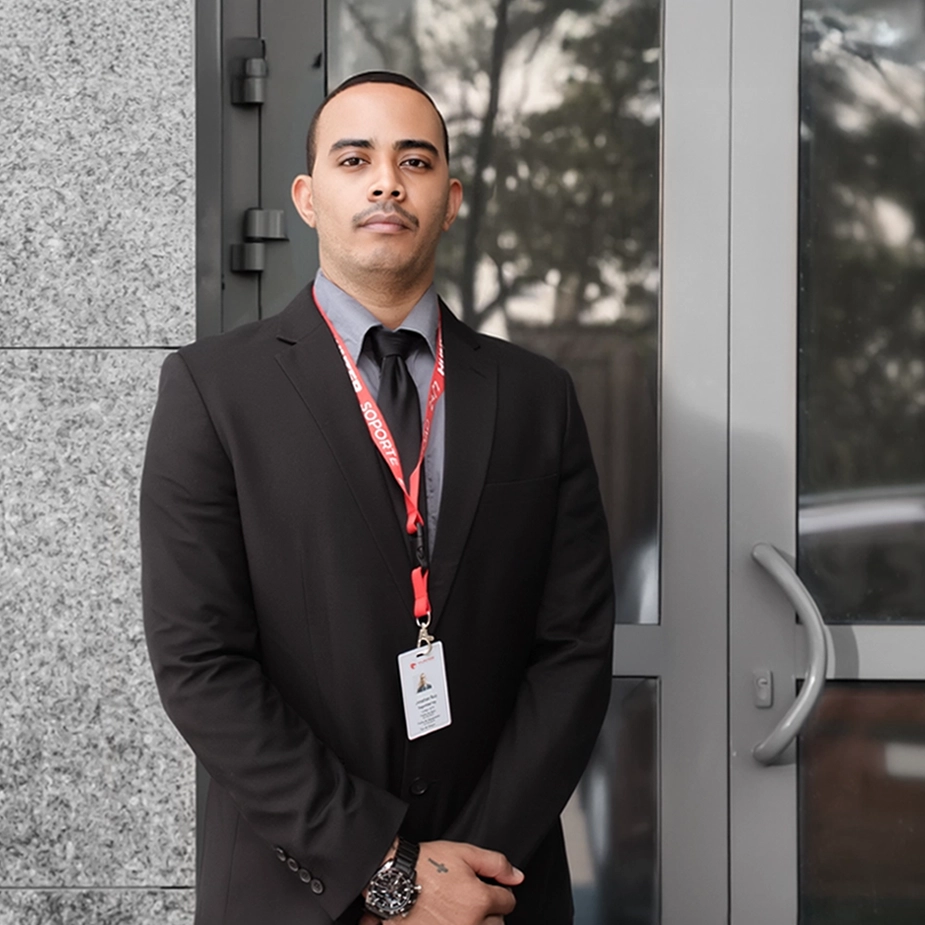 Man in a black suit with a red lanyard and ID badge standing in front of a glass door.
