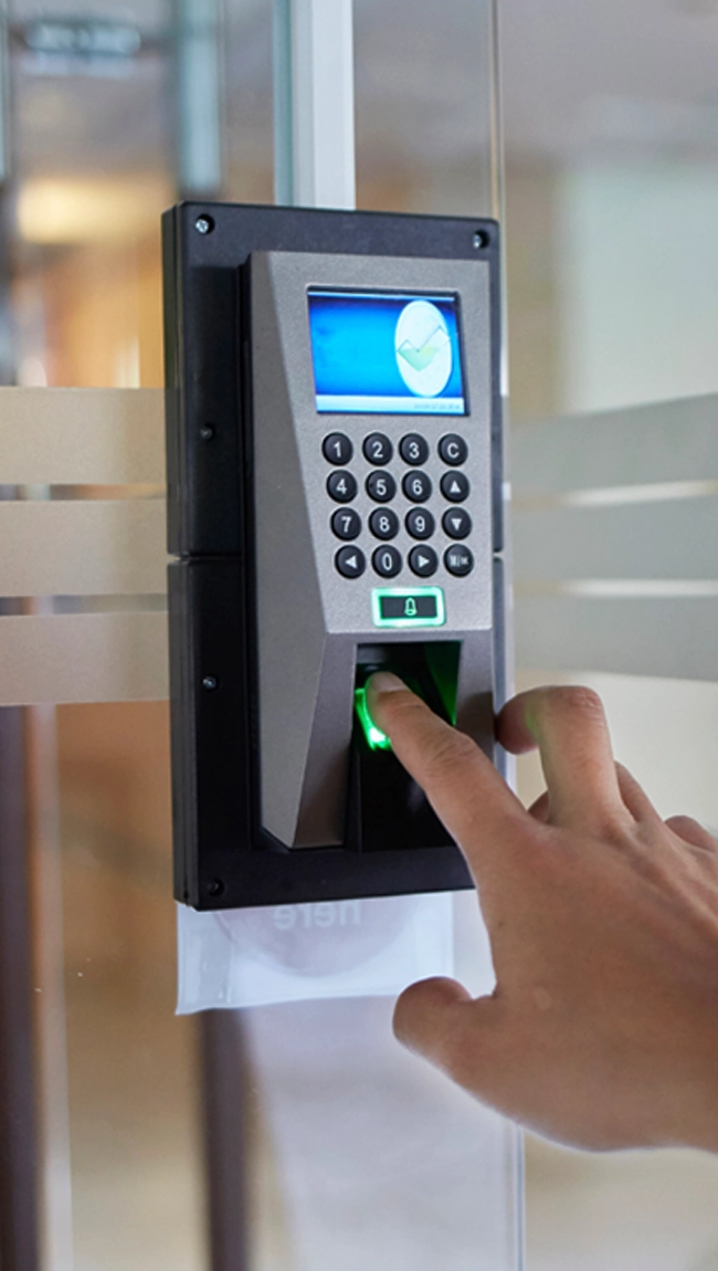 Hand placing finger on a fingerprint scanner integrated with a keypad access control system on a glass door.