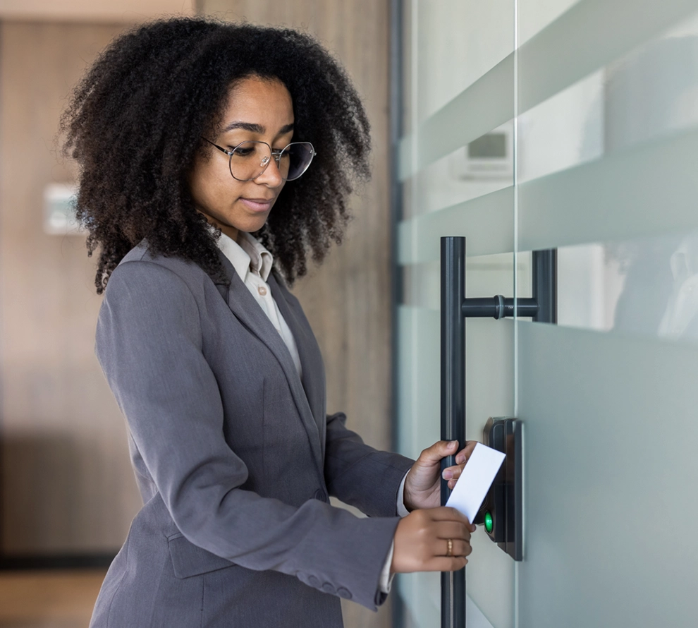 Woman in glasses and a gray blazer holding a white card to a security card reader on a glass door.