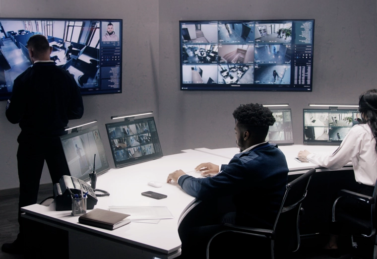 Security control room with three people monitoring multiple surveillance video screens.