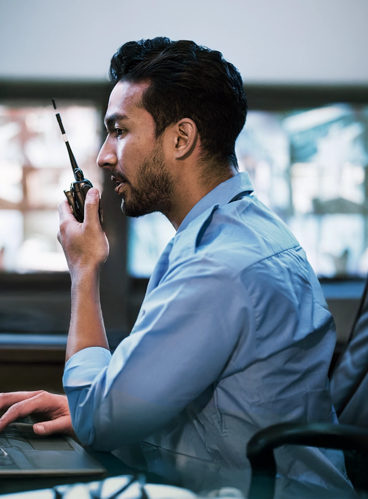 Man in a blue shirt using a walkie-talkie while working on a laptop indoors with blurred windows in the background.