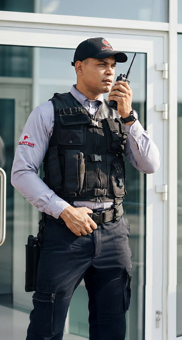 Security guard in uniform using a walkie-talkie outside a building with glass doors.
