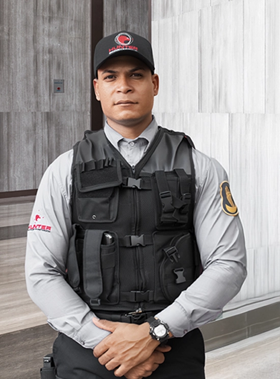 Security guard wearing a black tactical vest and cap standing with hands clasped inside a building.
