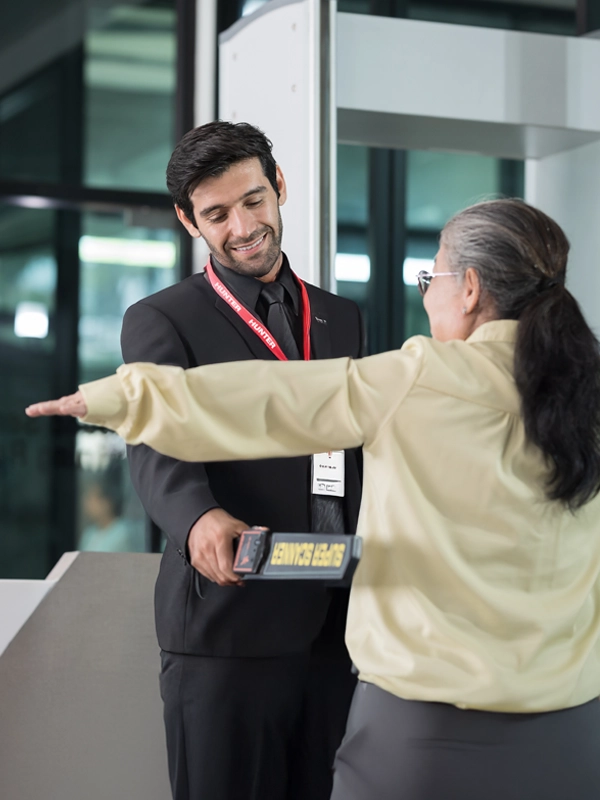 Security guard using a handheld metal detector to scan a woman with glasses and gray hair at an entrance checkpoint.