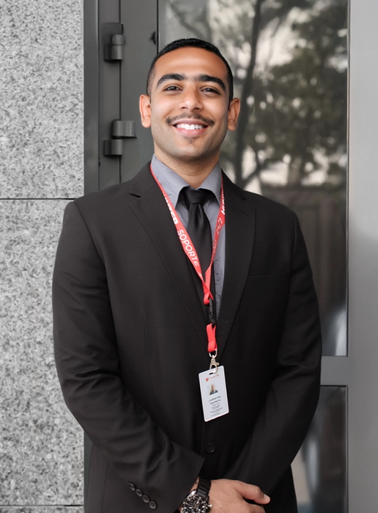 Smiling man in a black suit and tie wearing a red lanyard with an ID badge, standing against a stone wall and glass door.